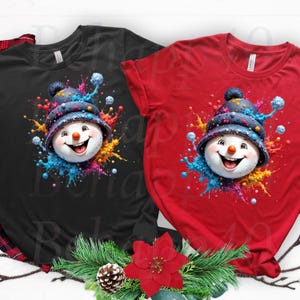 Op de afbeelding: Twee t-shirts met korte mouwen, één zwart en één rood, elk met een kleurrijke sneeuwpopafbeelding. De sneeuwpop heeft een blauwe hoed en is omgeven door verfspatten. Feestelijke vakantiekleding.
