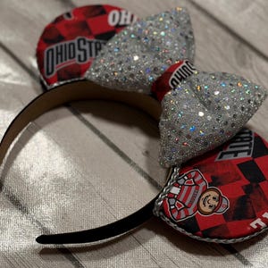May include: A black headband with decorative ears and a large, sparkly silver bow. The ears feature a red and black chequered pattern with the words "OHIO STATE" and a cartoon mascot.