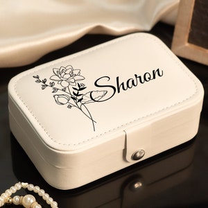 May include: A cream-colored jewelry box with a floral design and the name "Sharon" in black script. The box is rectangular with rounded edges and a silver clasp. A pearl necklace is in the foreground.
