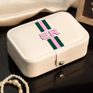 May include: A rectangular, off-white jewelry box with a pink and green stripe design and the letters "ER". The box is closed with a silver clasp. A pearl necklace is in the foreground.