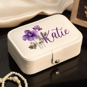 May include: Cream-colored rectangular jewelry box with a purple floral design and the name "Katie" in purple script. The box has a silver clasp and a stitched border.