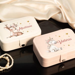 May include: Two rectangular jewelry boxes, one cream and one pink, with illustrations of a deer and a unicorn. The cream box has the name "Madison" and the pink box has the name "Victoria". Both boxes have gold star accents.