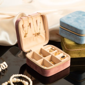 May include: A pink velvet jewelry box with a zipper closure. The box is open and has compartments for rings, earrings, and necklaces. There are three gold necklaces with charms inside the box. The box is sitting on a black surface.