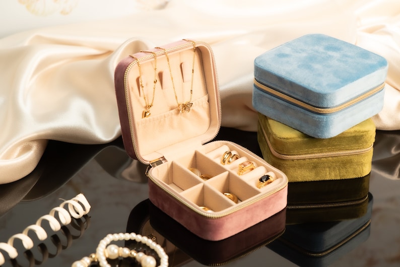 May include: A pink velvet jewelry box with a zipper closure. The box is open and has compartments for rings, earrings, and necklaces. There are three gold necklaces with charms inside the box. The box is sitting on a black surface.