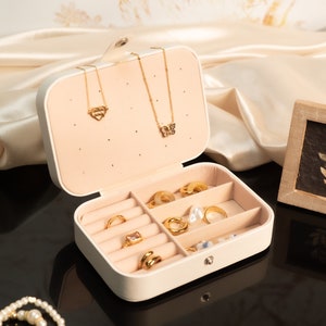 May include: A cream-colored jewelry box with a variety of gold rings, necklaces, and earrings. The box is open, revealing compartments filled with rings of different designs. Two gold necklaces hang from the top. A small framed botanical print sits nearby.