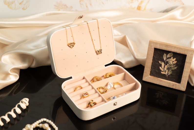 May include: A cream-colored jewelry box with a variety of gold rings, necklaces, and earrings. The box is open, revealing compartments filled with rings of different designs. Two gold necklaces hang from the top. A small framed botanical print sits nearby.