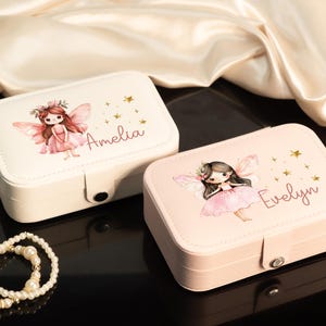 May include: Two rectangular jewelry boxes, one cream and one pink, each adorned with a fairy illustration and a name. The cream box has "Amelia" printed on it, and the pink box has "Evelyn". Gold star accents and a pearl bracelet are also visible.