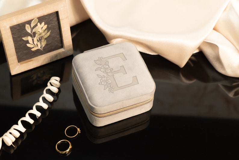 May include: A gray velvet jewelry box with a zipper closure. The box is decorated with an embroidered floral design and the letter "E".