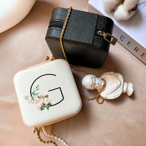 May include: Two square jewelry boxes with floral designs and gold chains. One box is white with a floral design and the letter "G" in black. The other box is black.