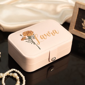 May include: A light pink jewelry box with the name "Laura" in gold script, adorned with a marigold floral design. The box is rectangular with a clasp closure. A framed botanical print and a pearl necklace are also visible.
