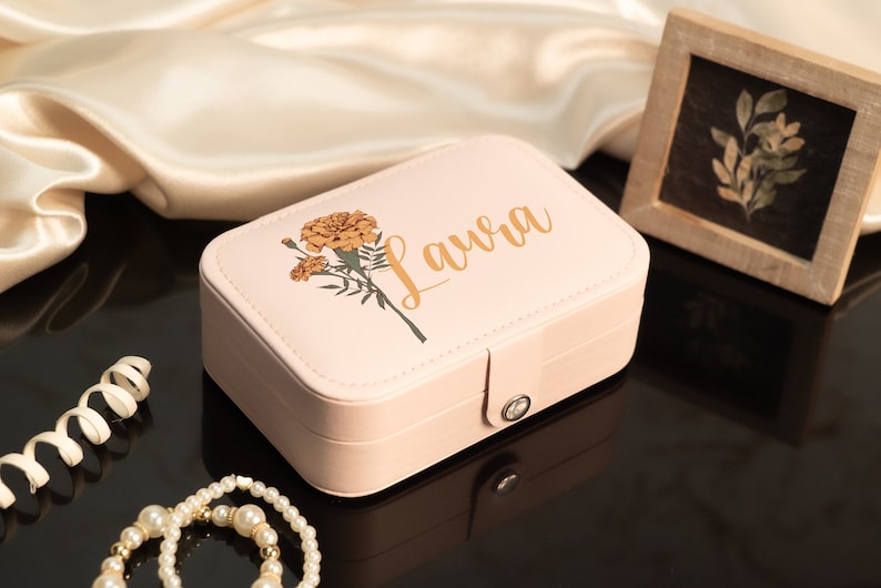 May include: A light pink jewelry box with the name "Laura" in gold script, adorned with a marigold floral design. The box is rectangular with a clasp closure. A framed botanical print and a pearl necklace are also visible.