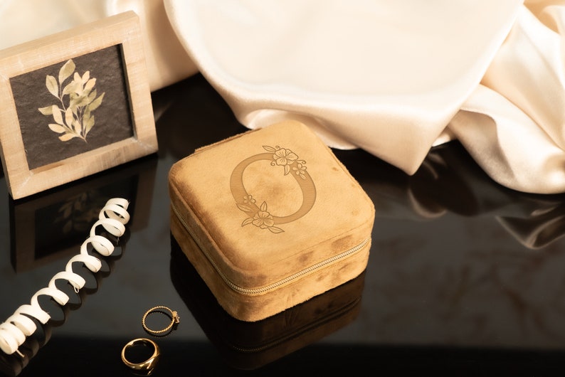 May include: A brown velvet jewelry box with a zipper closure. The box has a floral design with the letter "O" in the center. Two gold rings are on a black surface next to the box.