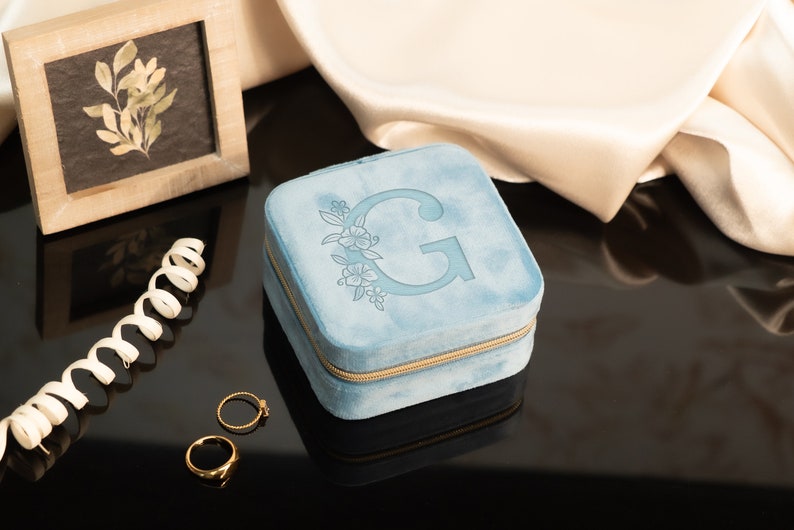 May include: A light blue velvet jewelry box with a gold zipper and a floral design with the letter "G" in the center.