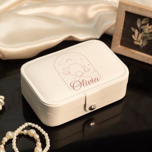 Op de afbeelding: Cr&egrave;mekleurige sieradendoos met een Weegschaal sterrenbeeld ontwerp en de naam "Olivia". De rechthoekige doos heeft een sluiting. Een parelketting en decoratief lint staan op de voorgrond. Een kleine ingelijste foto staat op de achtergrond.