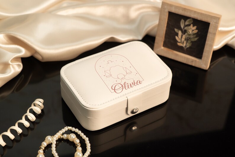 Op de afbeelding: Cr&egrave;mekleurige sieradendoos met een Weegschaal sterrenbeeld ontwerp en de naam "Olivia". De rechthoekige doos heeft een sluiting. Een parelketting en decoratief lint staan op de voorgrond. Een kleine ingelijste foto staat op de achtergrond.