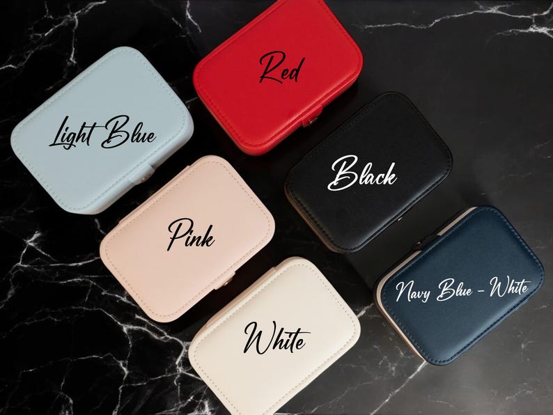 May include: A collection of small, rectangular jewelry cases in various colors: light blue, red, black, pink, white, and navy blue. Each case has a matching zipper and a label with the color name in cursive script.