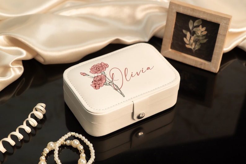 May include: A cream-colored rectangular jewelry box with a floral design and the name "Olivia" in a script font. The box is closed with a silver clasp. A pearl necklace and a small framed picture are also in the image.
