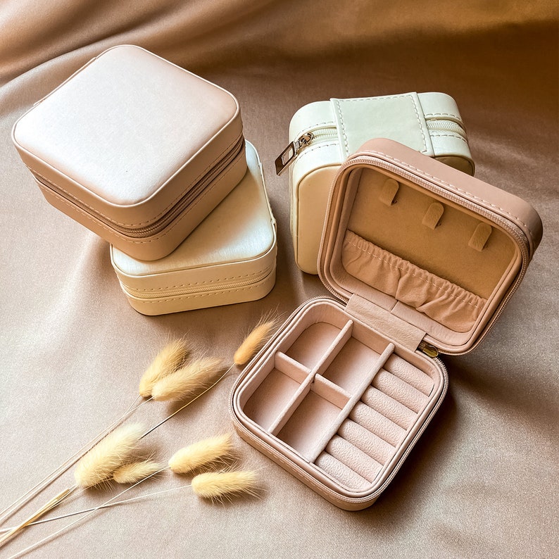 May include: Three square-shaped jewelry boxes with zippers. The boxes are beige and cream colored. The top box is open and has a compartment for rings and earrings.