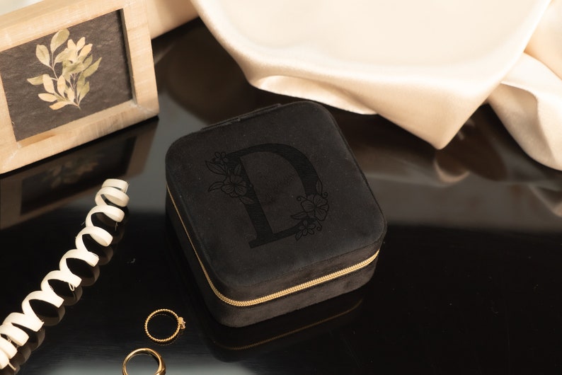 May include: Black velvet jewelry box with a gold zipper and the letter "D" embroidered on the top. The box is sitting on a black surface.