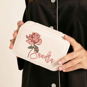 May include: A white jewelry case with a pink rose illustration and the name "Sandra" in a cursive font. The case is rectangular with a rounded top and is held by a person wearing a black pajama top.