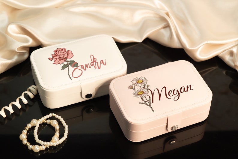 May include: Two rectangular jewelry boxes, one cream and one pink, are displayed. The cream box features a pink rose and the name "Sandra" in script. The pink box has daisies and the name "Megan". A pearl bracelet is in the foreground.