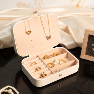Op de afbeelding: Een cr&egrave;mekleurige sieradendoos met een verscheidenheid aan gouden ringen en kettingen. De doos is open en onthult compartimenten gevuld met ringen van verschillende ontwerpen. Twee gouden kettingen met hart- en vlinderhangers hangen aan het deksel. Een kleine botanische print met lijst is ook zichtbaar.