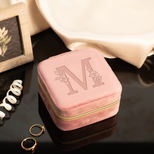 May include: A pink velvet jewelry box with a gold zipper and the letter "M" engraved on the top. The box is surrounded by a white satin fabric and two gold rings.
