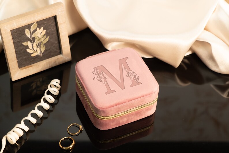 May include: A pink velvet jewelry box with a gold zipper and the letter "M" engraved on the top. The box is surrounded by a white satin fabric and two gold rings.
