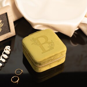 May include: A small, square, mustard yellow velvet jewelry box with a zipper closure. The box has a floral design and the letter "B" embossed on the top.