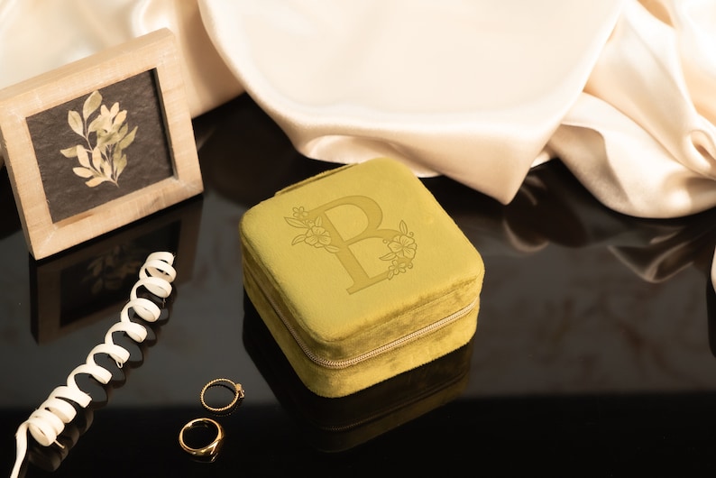 May include: A small, square, mustard yellow velvet jewelry box with a zipper closure. The box has a floral design and the letter "B" embossed on the top.