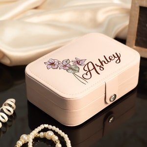 May include: A light pink jewelry box with a floral design and the name "Ashley" in a script font. The box is rectangular with a clasp closure. A pearl necklace and other jewelry are in the foreground.
