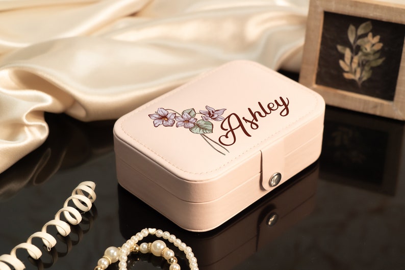 May include: A light pink jewelry box with a floral design and the name "Ashley" in a script font. The box is rectangular with a clasp closure. A pearl necklace and other jewelry are in the foreground.