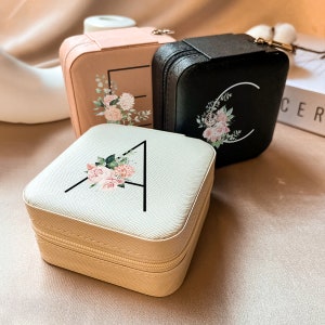 May include: Three small, square, zippered jewelry boxes with floral designs and a single letter on each. The boxes are beige, black, and pink.