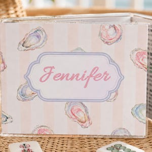 May include: A clear acrylic box with a pink and white striped design, adorned with watercolor oyster illustrations. The name "Jennifer" is displayed in pink script within a decorative frame. The box is open, revealing printed pages.