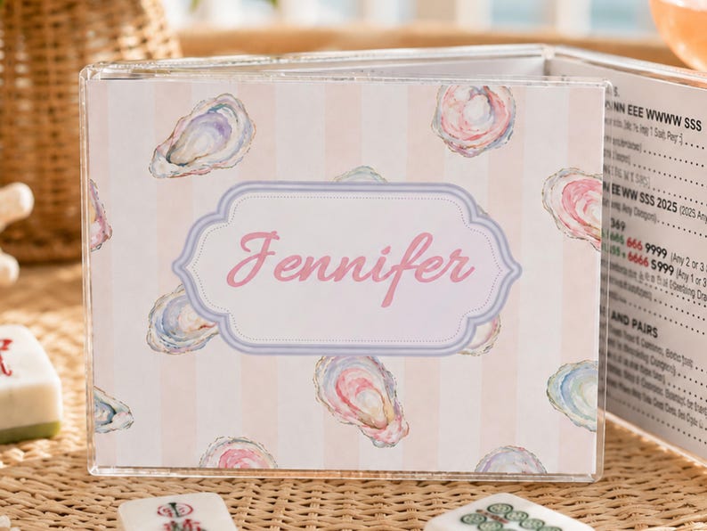 May include: A clear acrylic box with a pink and white striped design, adorned with watercolor oyster illustrations. The name "Jennifer" is displayed in pink script within a decorative frame. The box is open, revealing printed pages.