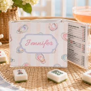 May include: A clear acrylic menu holder with a pink and white striped design, featuring watercolor oyster illustrations. The holder displays a menu with pricing and the name "Jennifer" in pink script. Several Mahjong tiles are in the foreground.