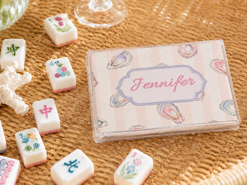 May include: A clear plastic case with the name "Jennifer" in pink script, surrounded by a light pink and white striped background with oyster illustrations. Scattered around are white and pink Mahjong tiles with various symbols and designs.