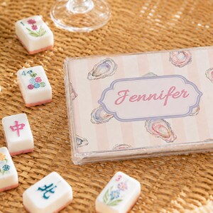 May include: A clear plastic case with the name "Jennifer" in pink script, surrounded by a light pink and white striped background with oyster illustrations. Scattered around are white and pink Mahjong tiles with various symbols and designs.