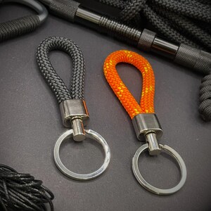 May include: Two keyrings with braided rope loops. One is black, the other is orange with yellow accents. Both have silver-coloured metal hardware and key rings. The background includes more black rope and metal hardware.