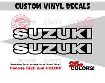 May include: Two white vinyl decals featuring the word SUZUKI in a bold, block-style font with a black outline. The text is presented in two horizontal rows against a light gray background, with additional text including CUSTOM VINYL DECALS.