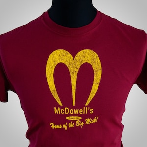 May include: A red t-shirt with a yellow stylized "M" logo and the text "McDowell's Queens, NY Home of the Big Mick!"