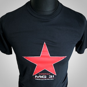May include: Black t-shirt featuring a large, distressed red star graphic. Below the star, the text reads "FIREFOX MIG 31 MIKOYAN-GUREVICH" in white. The shirt is displayed on a mannequin.