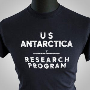 May include: A black t-shirt with white text that reads "US ANTARCTICA RESEARCH PROGRAM." Below the text is "UNITED STATES NATIONAL SCIENCE INSTITUTE STATION 4." The shirt is displayed on a black mannequin.