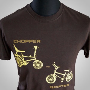 May include: A brown t-shirt with a gold graphic of two bicycles. The text "CHOPPER" is above the first bicycle and the text "GRIFTER" is below the second bicycle. The text "VS" is between the two bicycles.