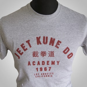 Puede incluir: Camiseta gris con un estampado gráfico rojo que dice "Jeet Kune Do Academy 1967 Los Angeles California" con caracteres chinos en el medio.