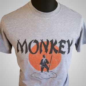 Könnte beinhalten: Ein graues T-Shirt mit einer schwarz-weißen Grafik einer Person, die auf einer Wolke mit einer roten Sonne dahinter steht. Der Text "MONKEY" ist in Schwarz über der Grafik gedruckt.