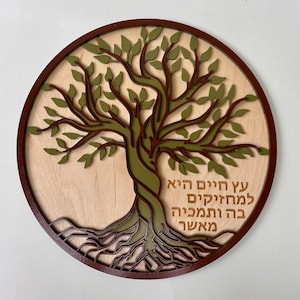 May include: Round wooden wall art featuring a tree design with a dark brown frame. The tree has green leaves and a brown trunk and roots. The background is a light wood color, and text is visible on the right side of the tree.