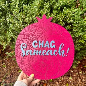 May include: A vibrant pink pomegranate-shaped wooden sign with intricate etched patterns. The sign features the words "CHAG Sameach!" in light blue, celebrating a Jewish holiday. The background is a lush green bush.