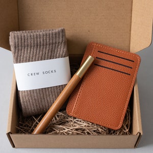 May include: A brown cardboard box with a brown pair of socks, a brown leather card holder, and a wooden pen with a gold top. The socks have a white label that says "CREW SOCKS".
