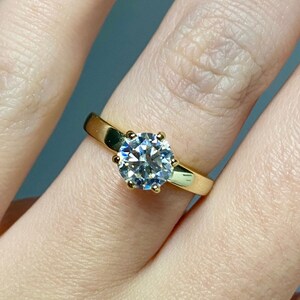 May include: A gold ring featuring a large, round, clear gemstone. The ring has a simple, elegant design with a smooth, polished band. The gemstone is set in a classic four-prong setting, highlighting its sparkle.